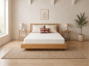 A minimalist wooden platform bed with a white textured duvet and matching pillows, styled in a bright, modern bedroom with a woven jute rug, wooden floor, and side tables.