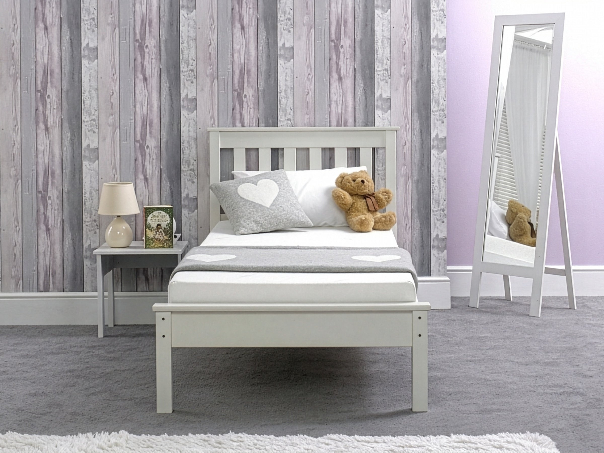 A child's bedroom featuring a white bed frame with a memory foam mattress, a grey rug, and a teddy bear on the bed. The wall behind the bed features a wood panel design in grey tones.