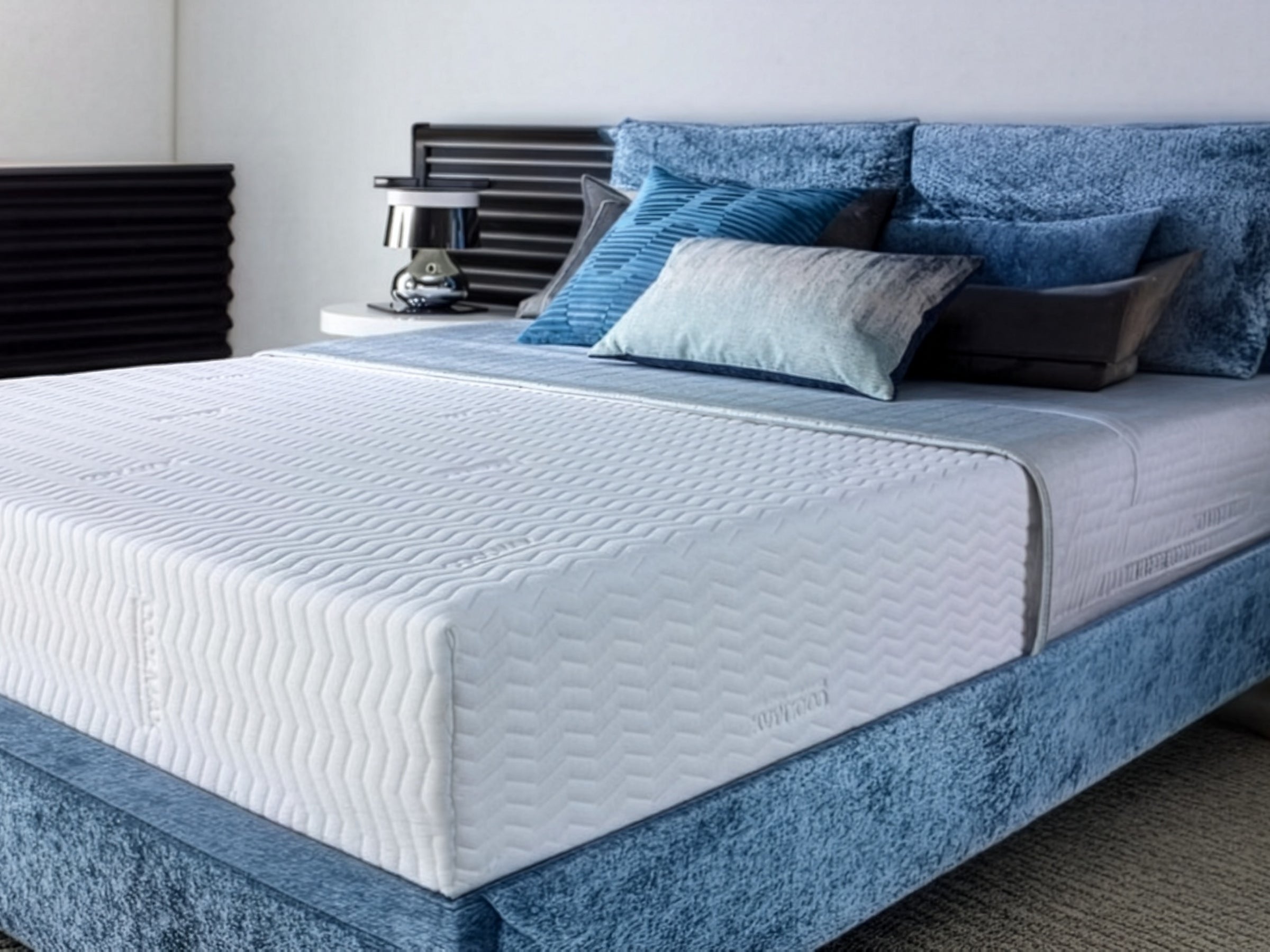 A white quilted mattress cover on a bed with blue bedding, in a room with a window and curtains.