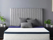 A memory foam mattress topper on a bed, with a grey headboard and dark furniture around the bed. Plants are visible beside the bed, and the room has a grey wall.