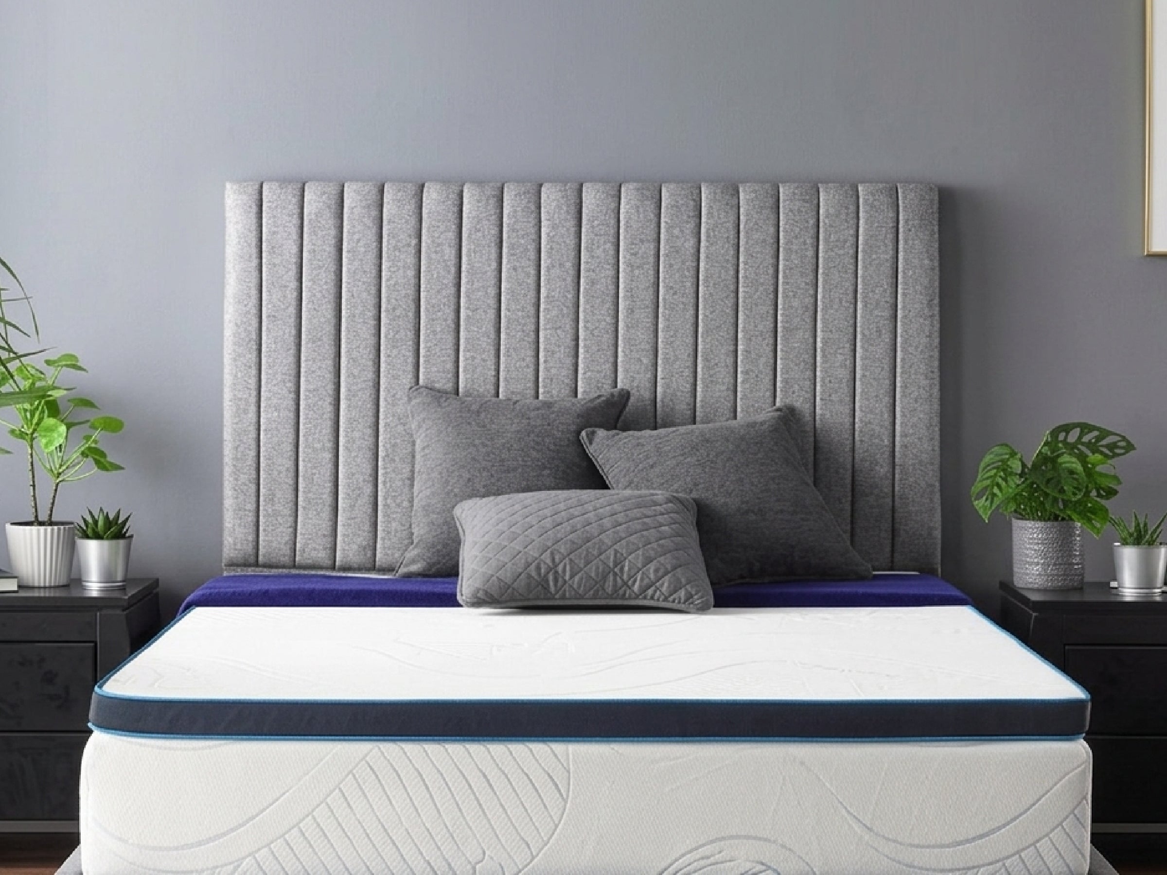 A memory foam mattress topper on a bed, with a grey headboard and dark furniture around the bed. Plants are visible beside the bed, and the room has a grey wall.