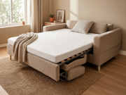 A beige upholstered sleeper sofa in a bright room, shown with a white mattress pulled out and a matching throw blanket draped over the side.
